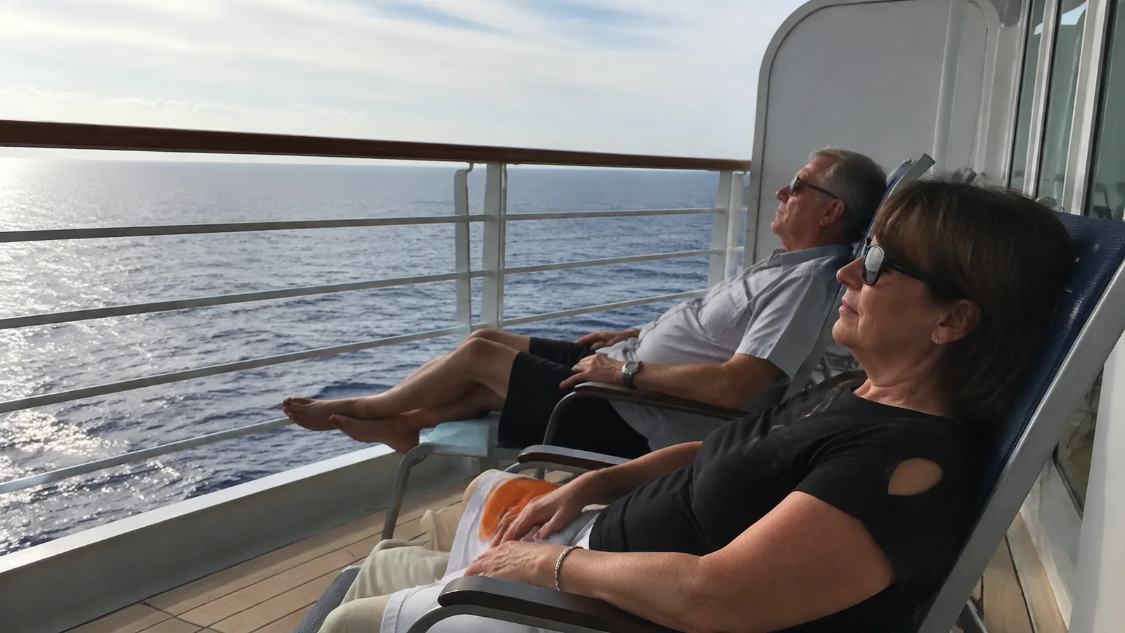 Couple relaxes in balcony chairs overlooking the sunlit ocean.