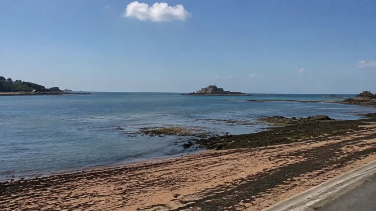 Calm coastal bay with exposed rocks and a distant offshore fort.
