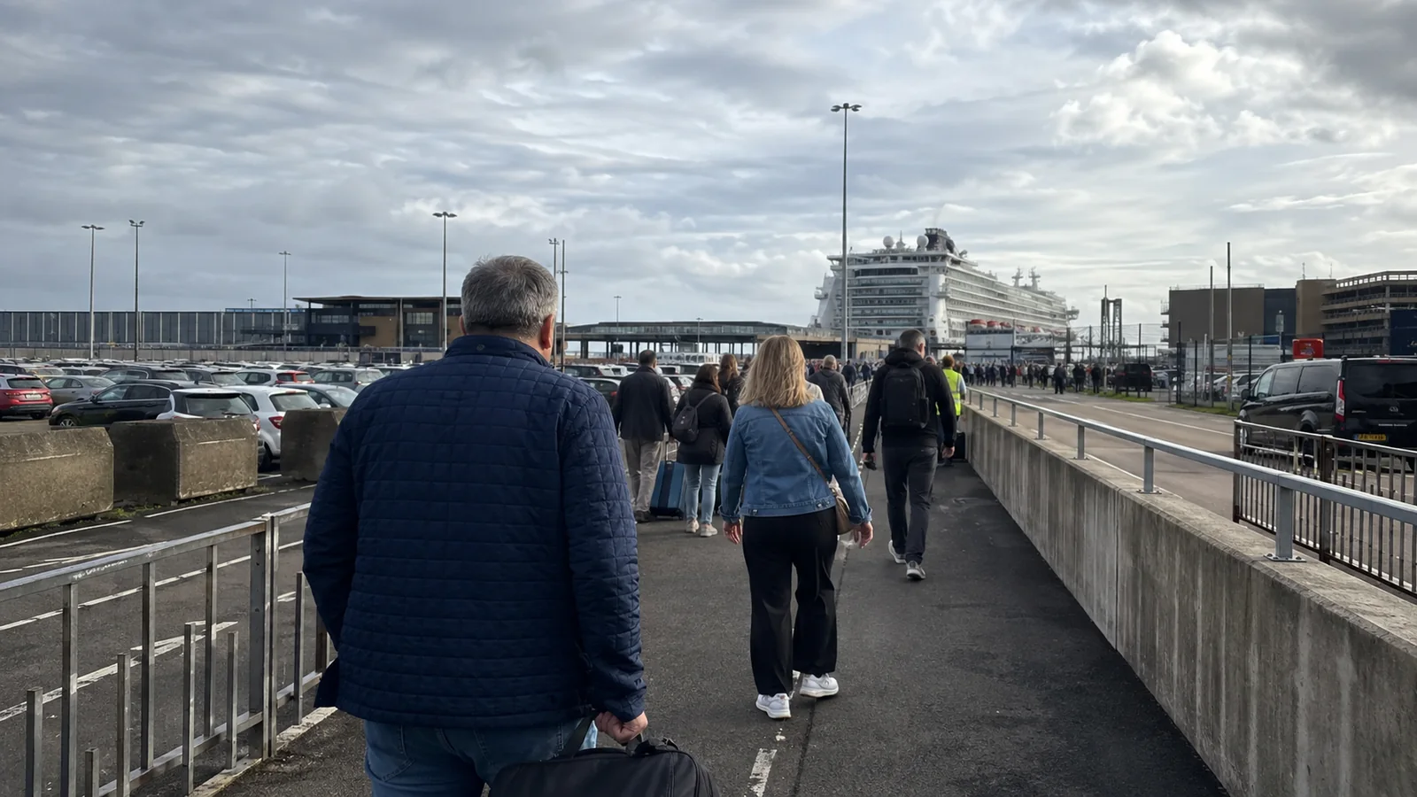 Passengers walking toward a cruise terminal beside a car park.