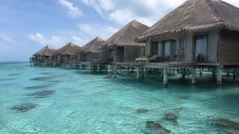 Row of thatched overwater villas above a clear turquoise lagoon.