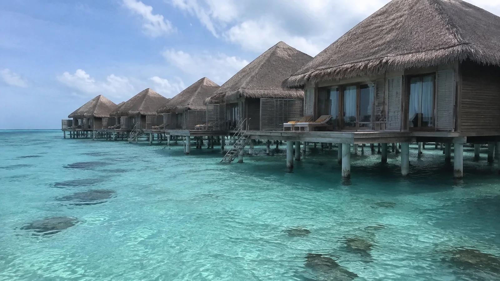 Row of thatched overwater villas above a clear turquoise lagoon.