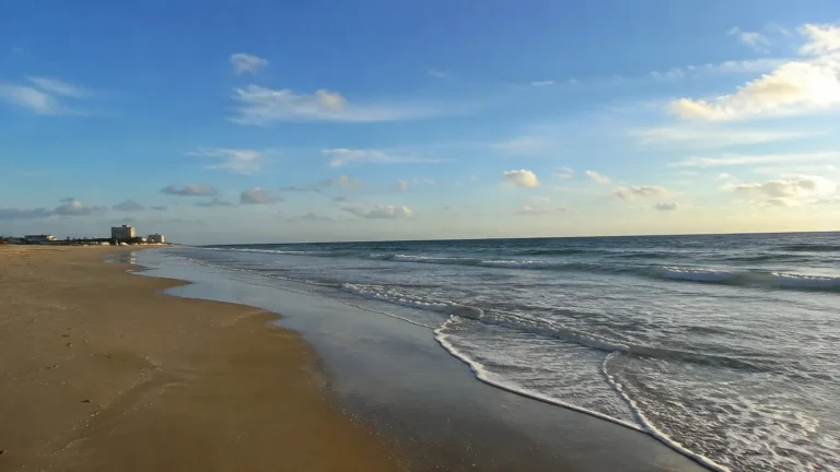 Gentle waves meeting a broad sandy beach beneath a lightly clouded sky.