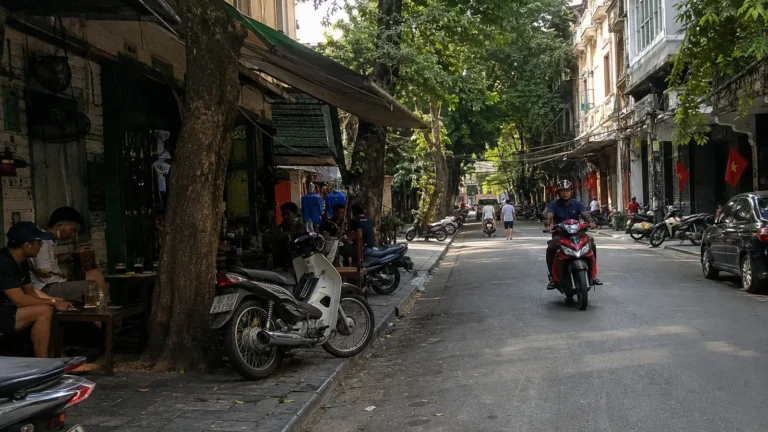 Motorbikes passing cafes and shops along a shaded city street.