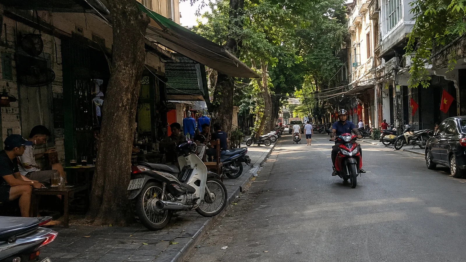 Motorbikes passing cafes and shops along a shaded city street.