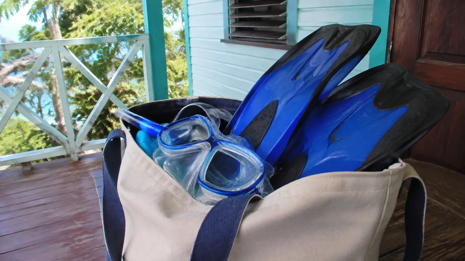 Blue snorkeling fins and a mask packed into a canvas tote on a porch.
