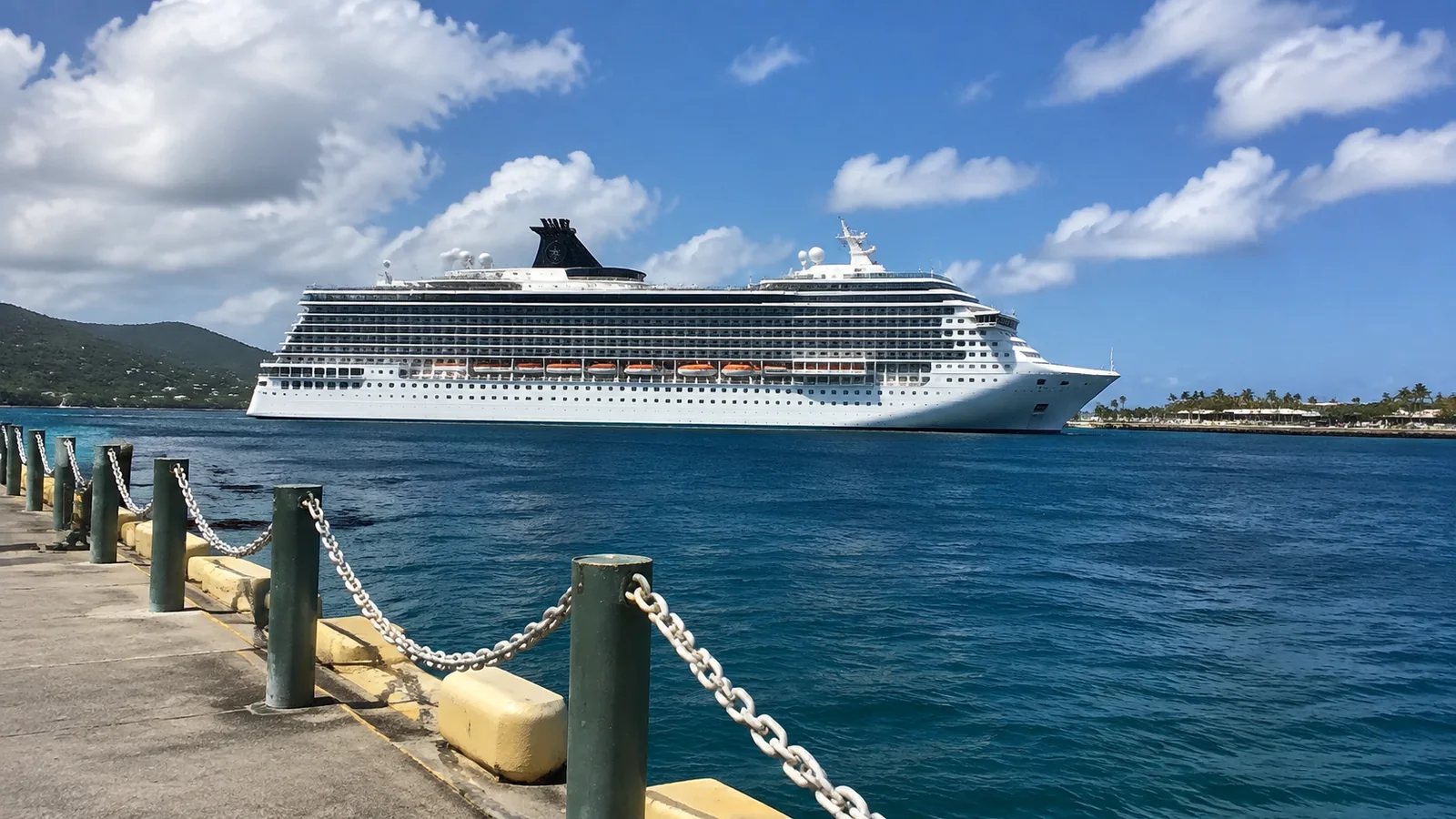 Large white cruise ship beside a tropical waterfront under scattered clouds.