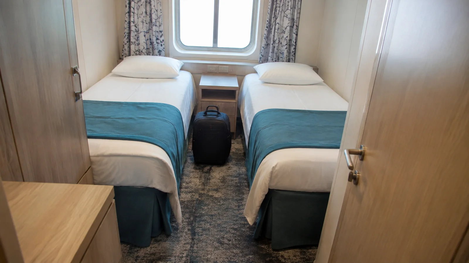 Compact ship cabin with twin beds, turquoise bed runners, and a small window.