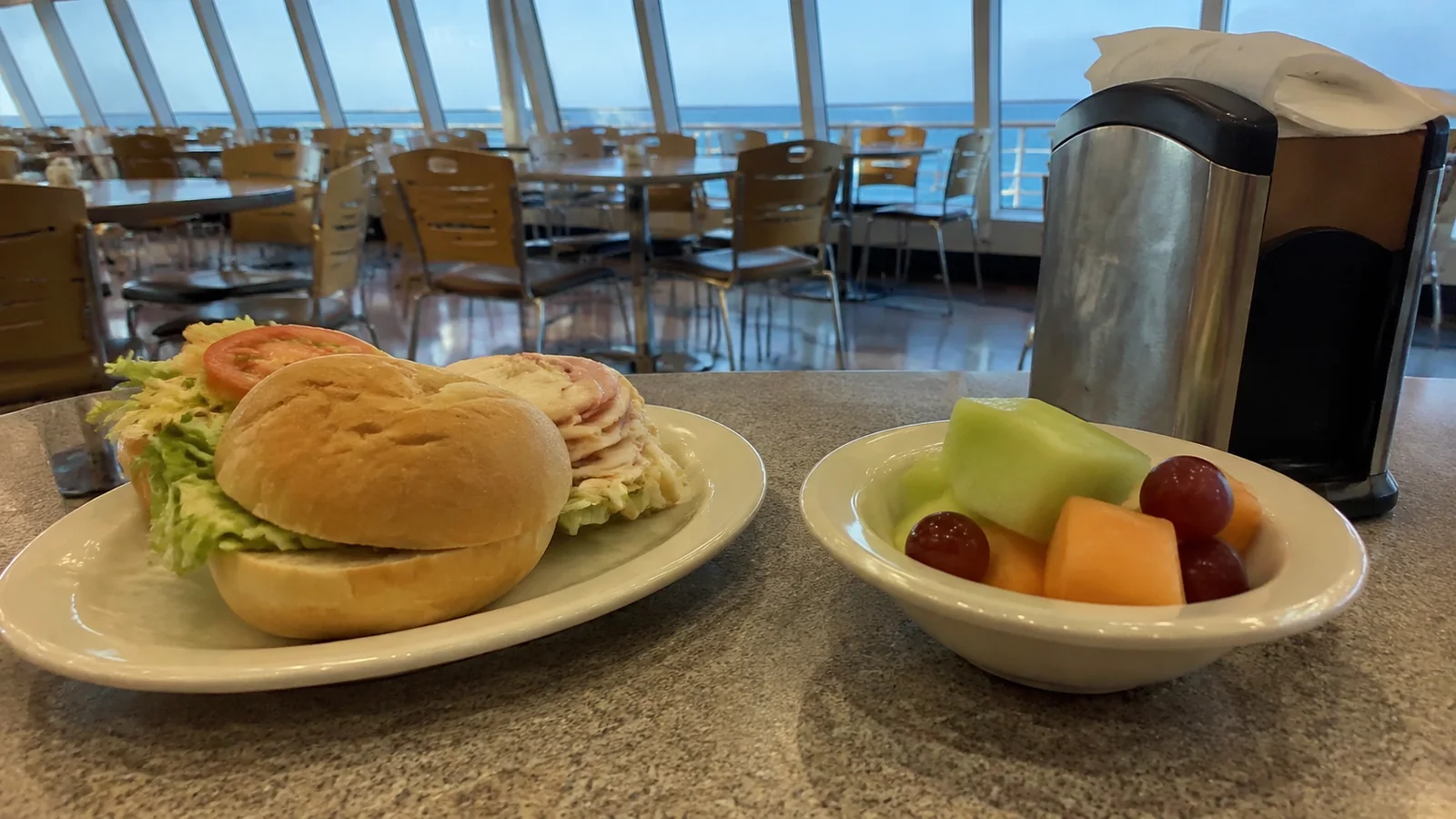 Sandwich and bowl of cut fruit on a cafeteria table aboard a ship.