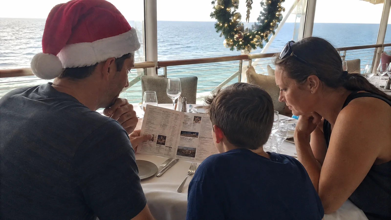 Family seated at a cruise ship dining table reviewing a paper while a man wears a Santa hat