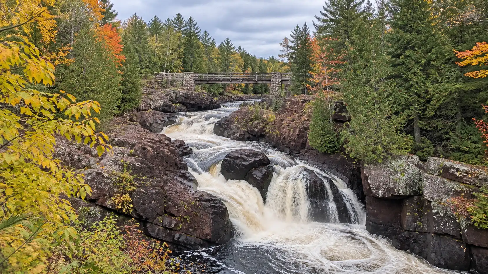 Copper Falls flows through a forested reddish-brown gorge beside a rustic bridge.