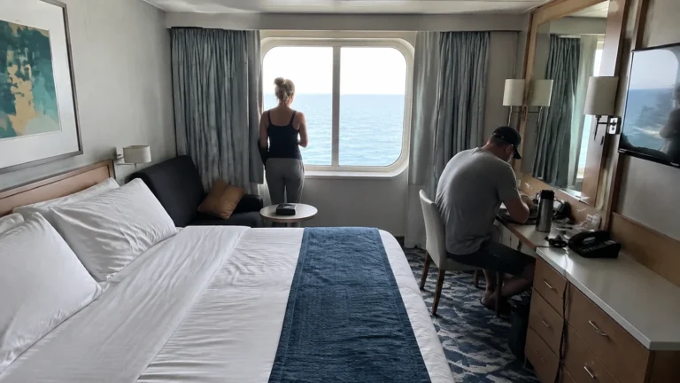 Two passengers settling into an ocean-view cabin with a bed, sofa and desk.
