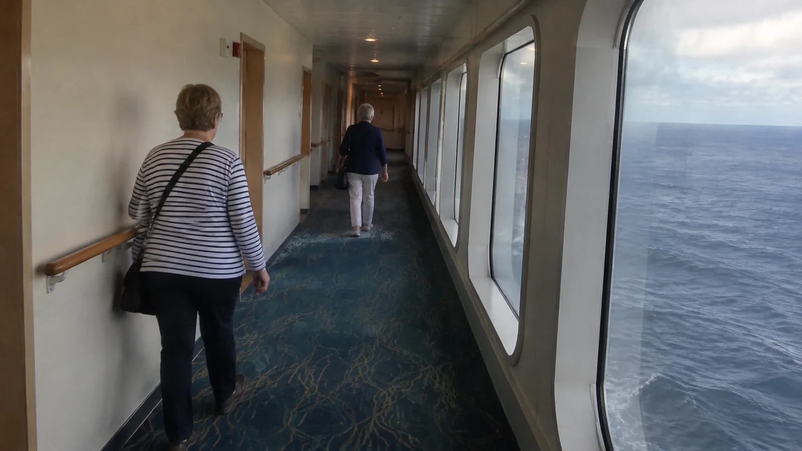 Passengers walking down an enclosed corridor with large ocean-view windows.