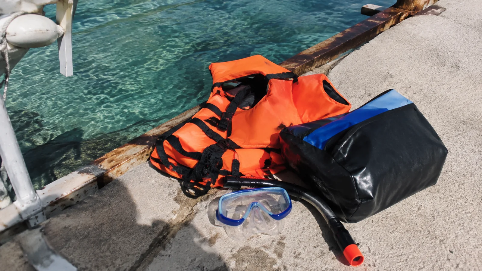 Life jackets, snorkel mask and dry bag beside clear water at a dock.