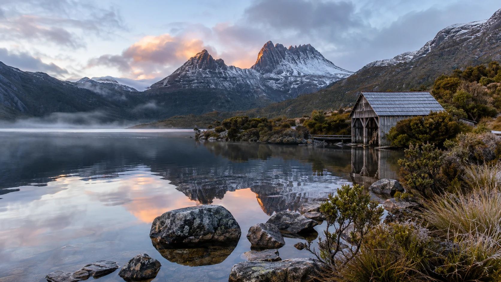 Snow-dusted Cradle Mountain and the Dove Lake boatshed reflect in calm Tasmanian water at dawn.