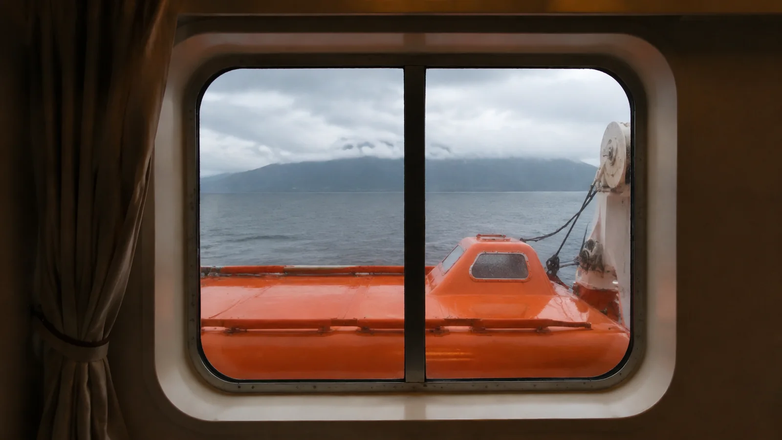 Orange lifeboat partially blocking the view through a cabin window.