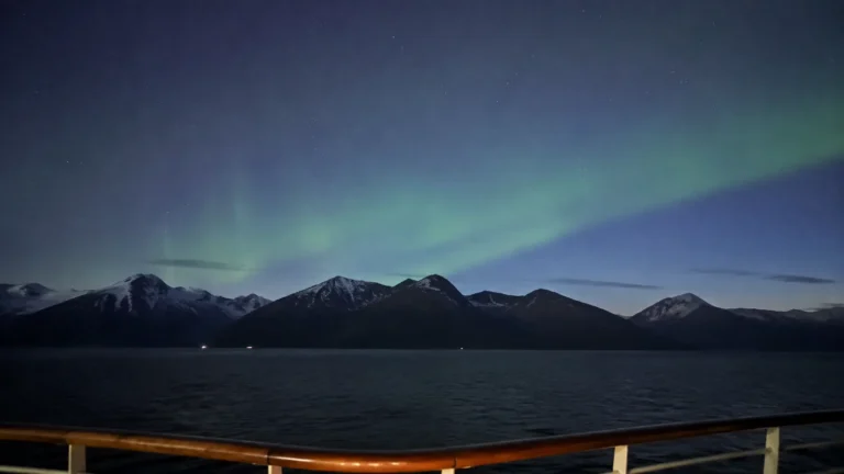 Green northern lights above a dark mountain shoreline viewed from a ship.