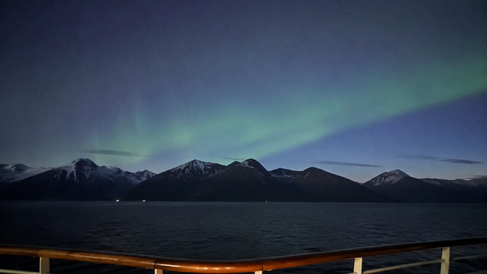 Green northern lights above a dark mountain shoreline viewed from a ship.