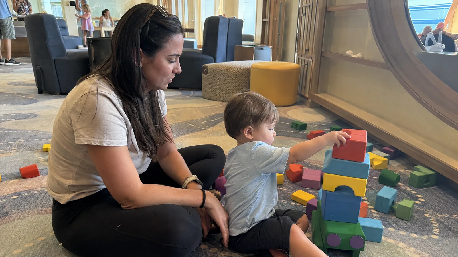 Woman sitting beside a young child building a tower of colorful blocks.