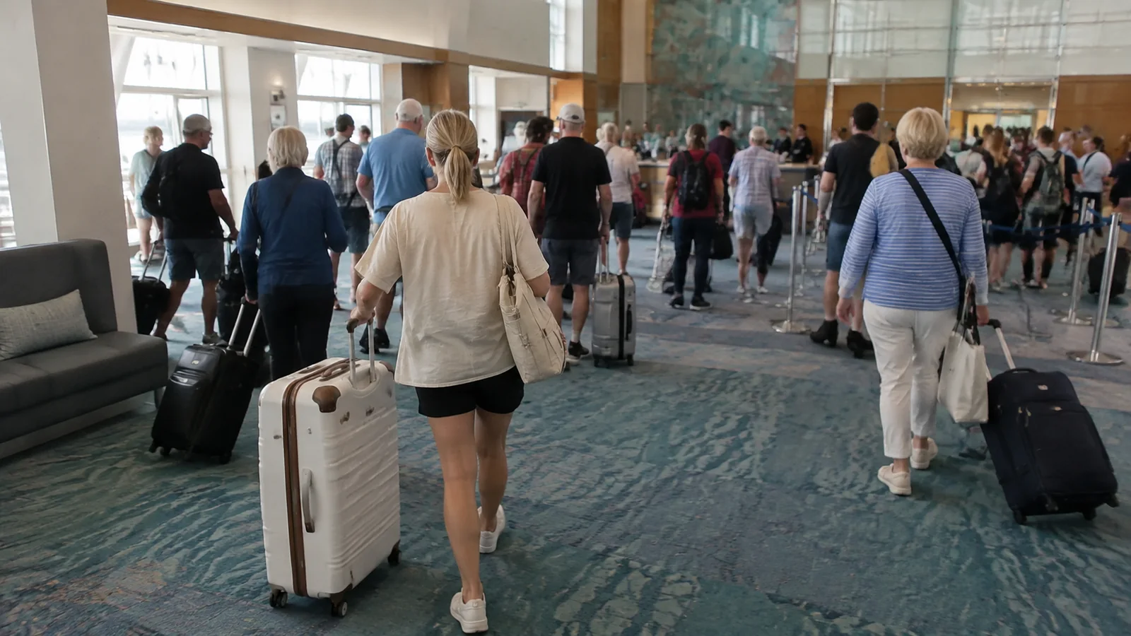 Travelers pull suitcases through a busy terminal toward queue barriers.