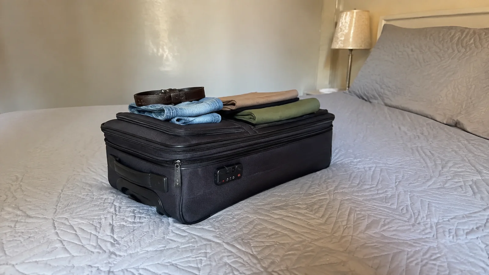 Closed black suitcase topped with neatly folded clothes on a bed.
