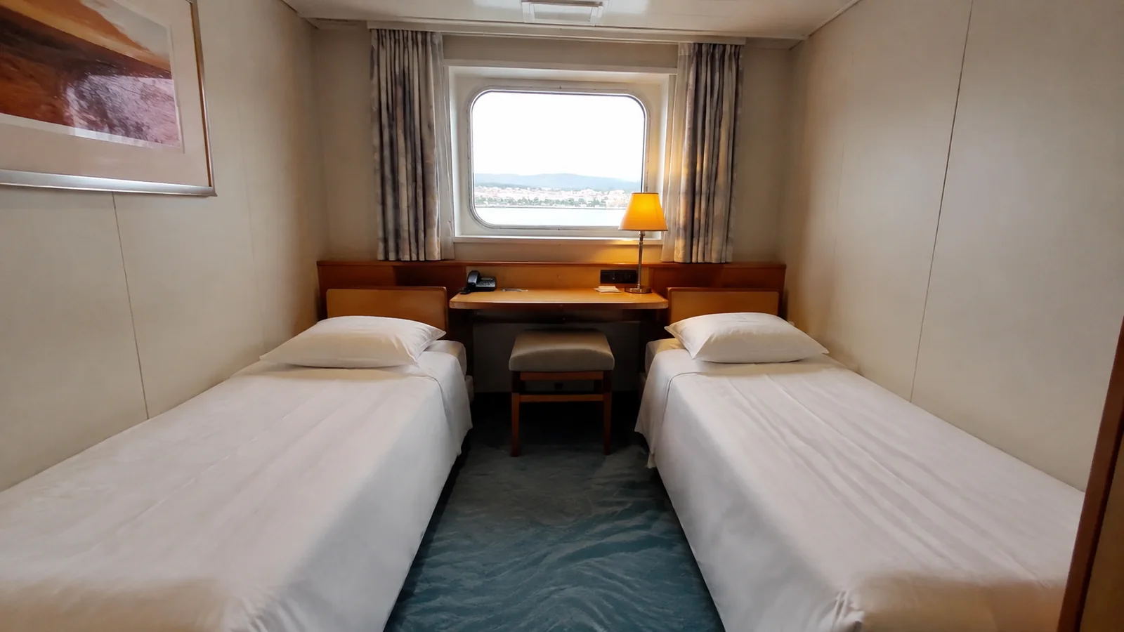Narrow ship cabin with twin beds and a window above a small desk.