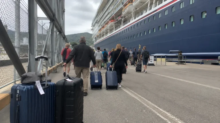 Travelers rolling suitcases along a pier beside a docked cruise ship