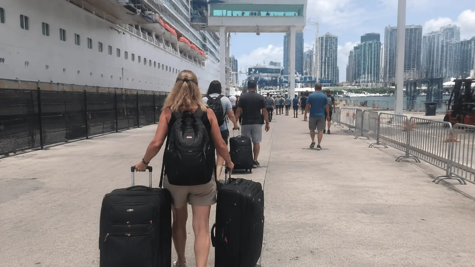 Woman pulling two suitcases along a cruise pier toward a city skyline