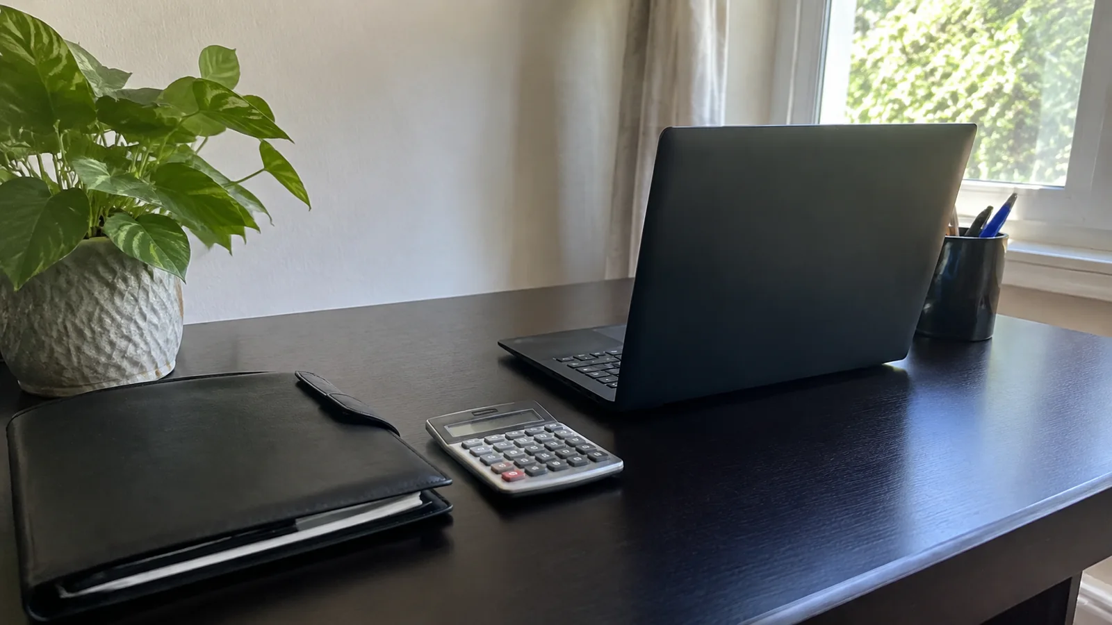 Open laptop, calculator and dark folder sit on a desk beside a plant.
