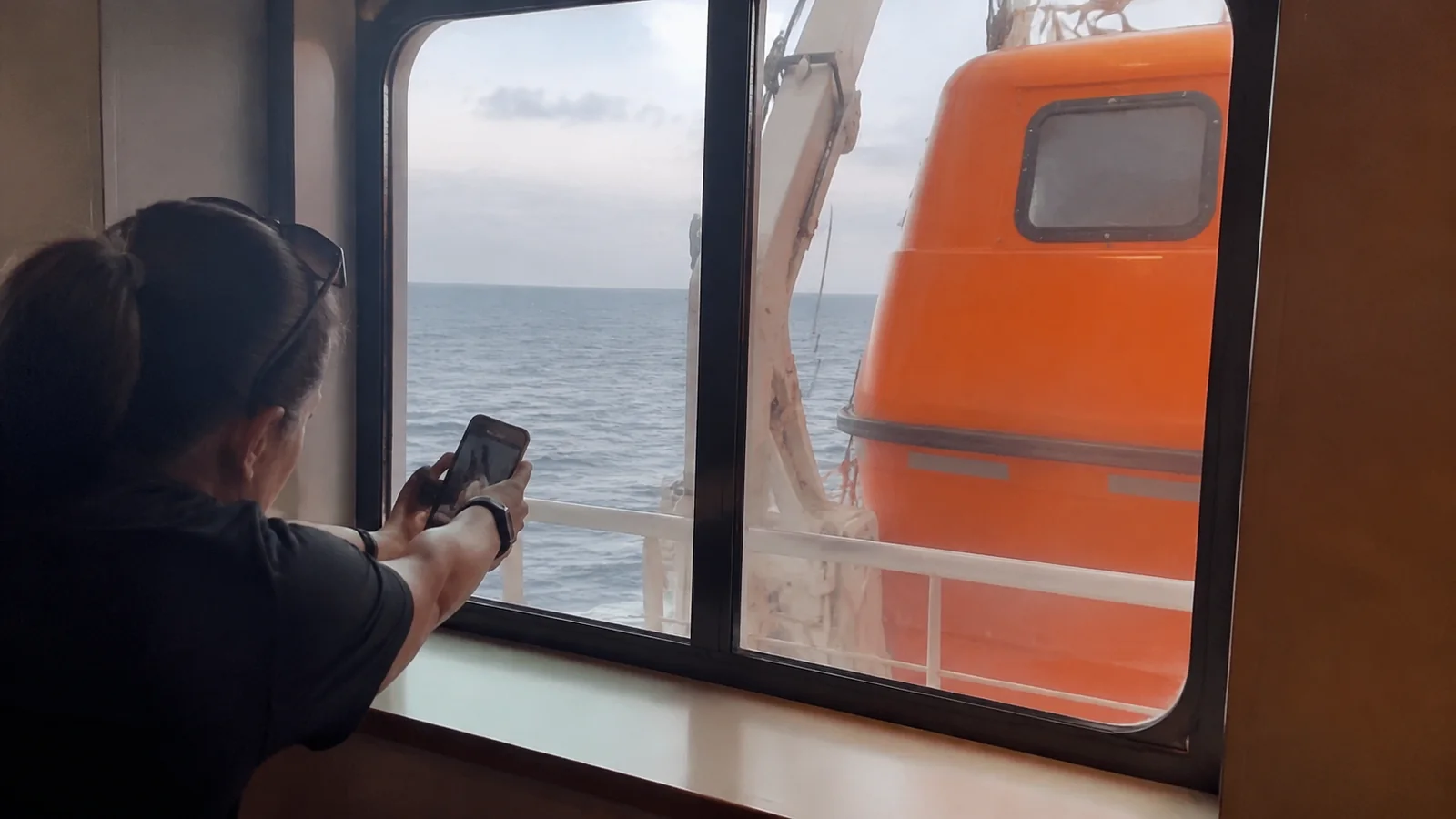 Passenger using a phone beside a window partly blocked by a lifeboat.