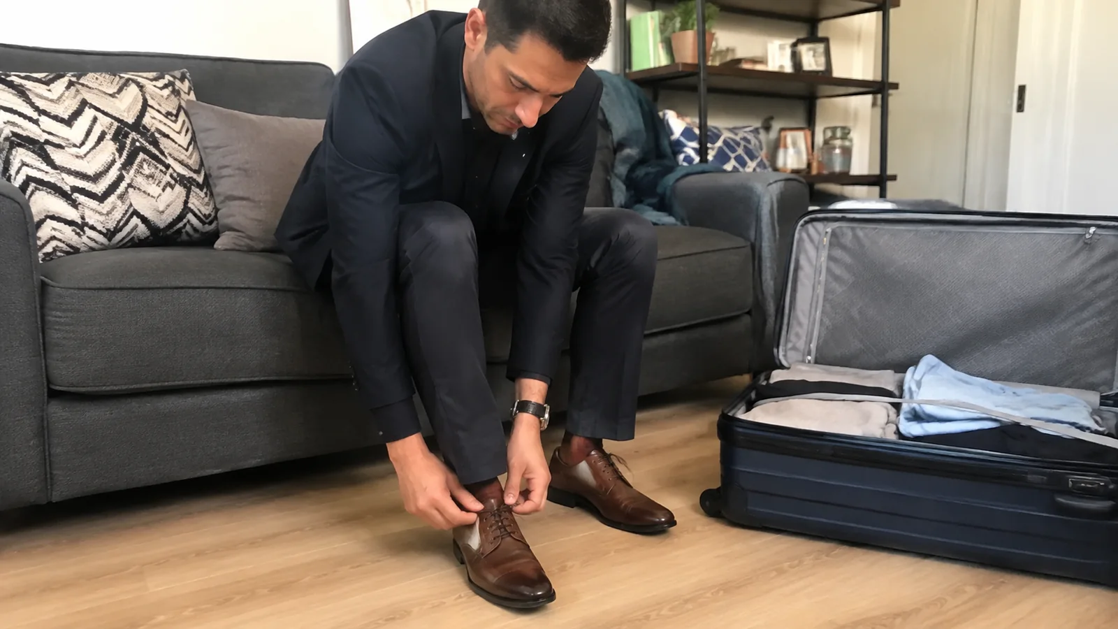 Man tying leather shoes while wearing a dark suit beside packed luggage.