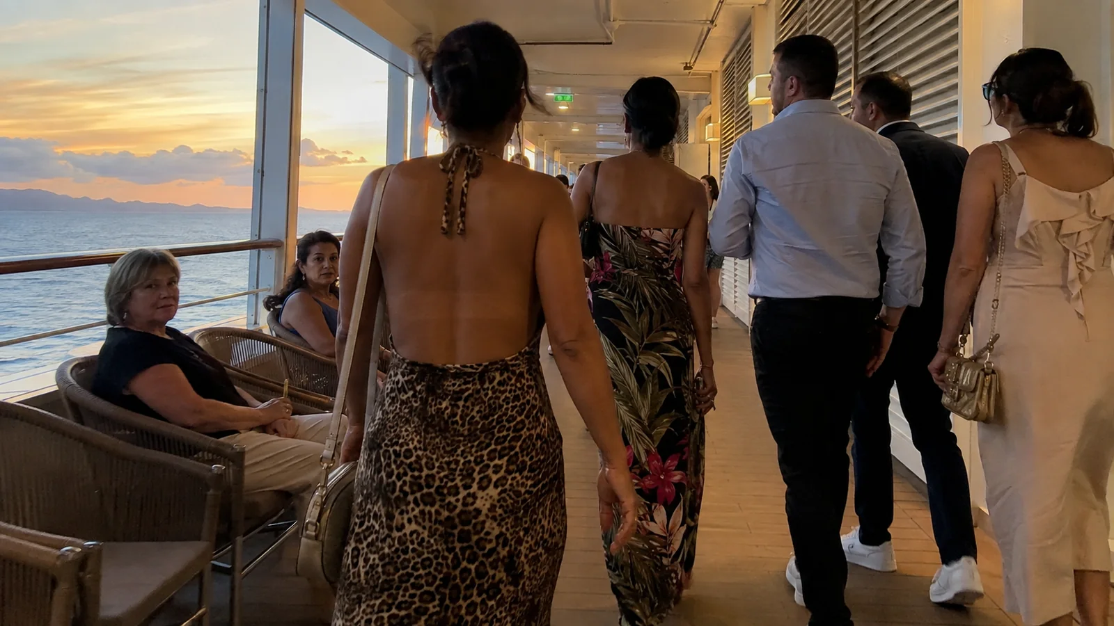 Passengers in evening clothes walking through an ocean-view lounge at sunset.