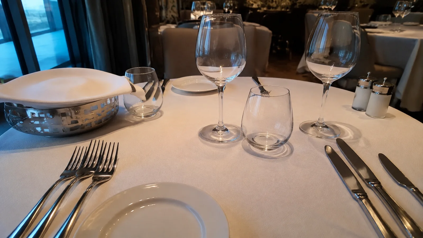 Restaurant table set with plates, forks, knives, wine glasses, and a bread basket.