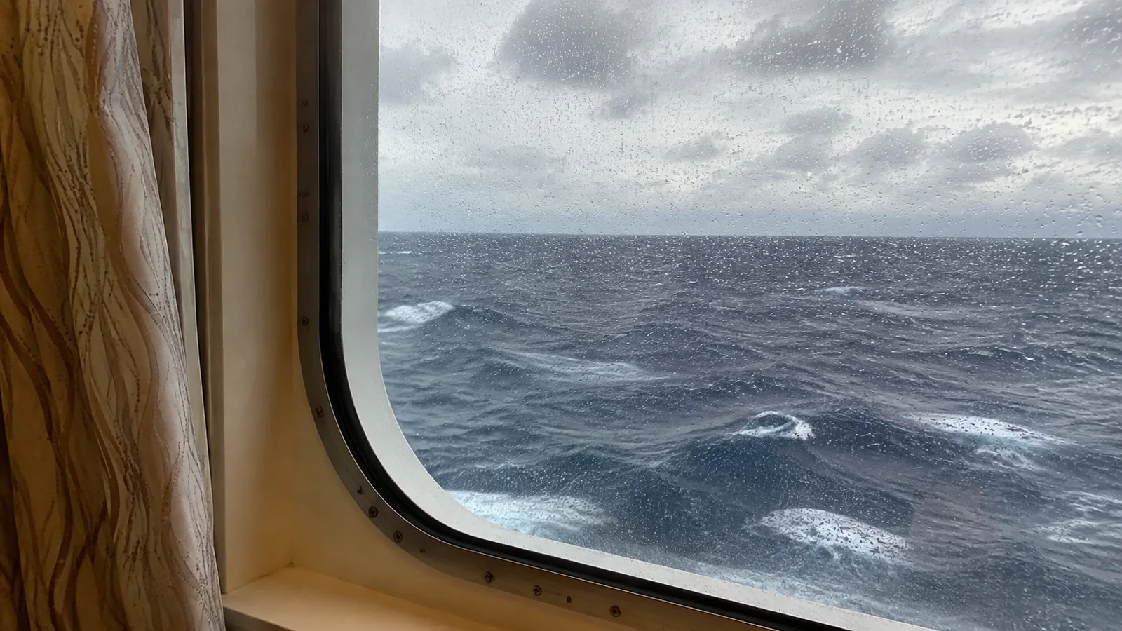 Rain-speckled window frames dark water and white ocean waves.