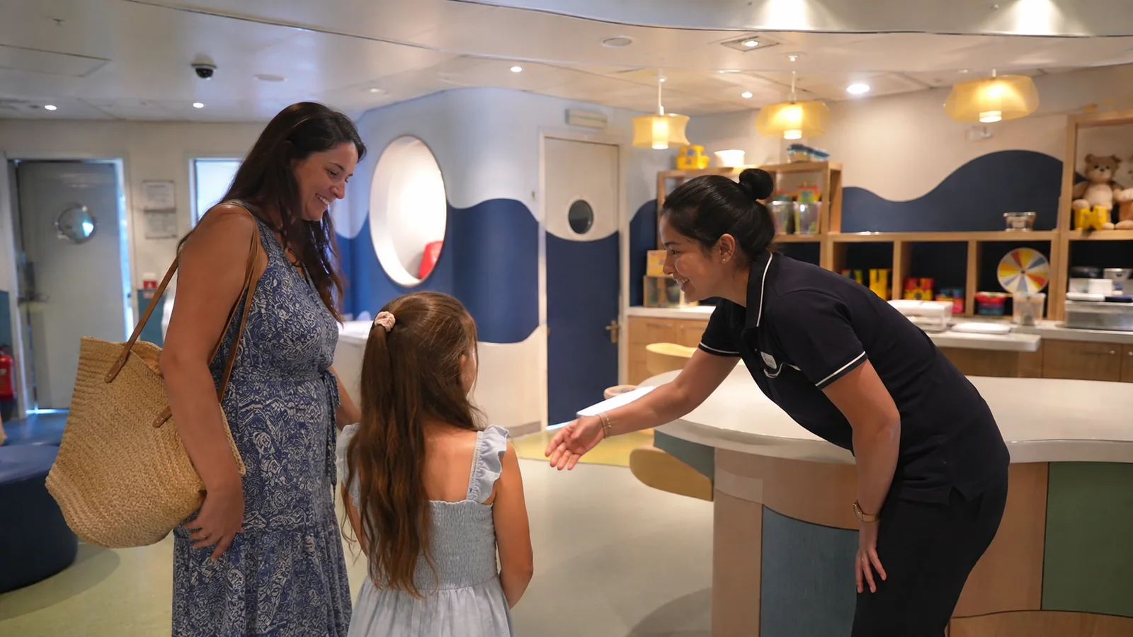 Staff member greeting a woman and child at a ship activity room entrance.