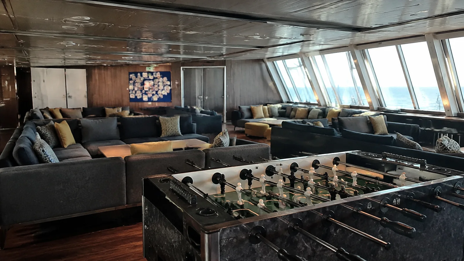 Ship lounge with sofas, a foosball table, and slanted ocean-facing windows.