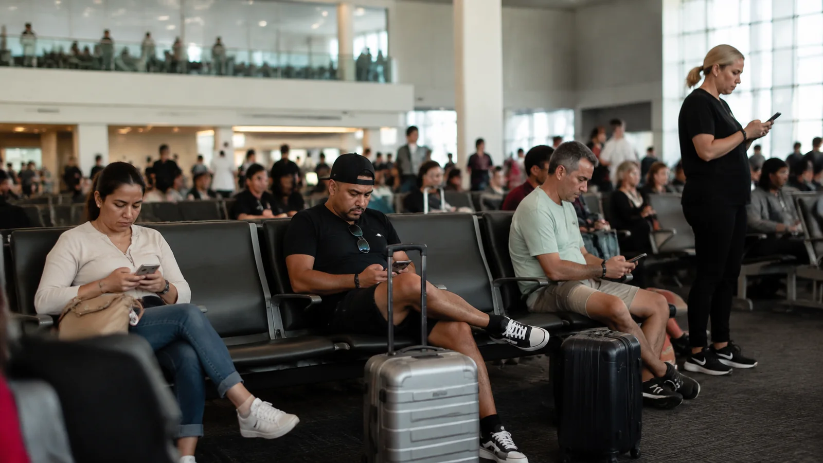 Travelers sit with luggage and phones in a busy terminal.