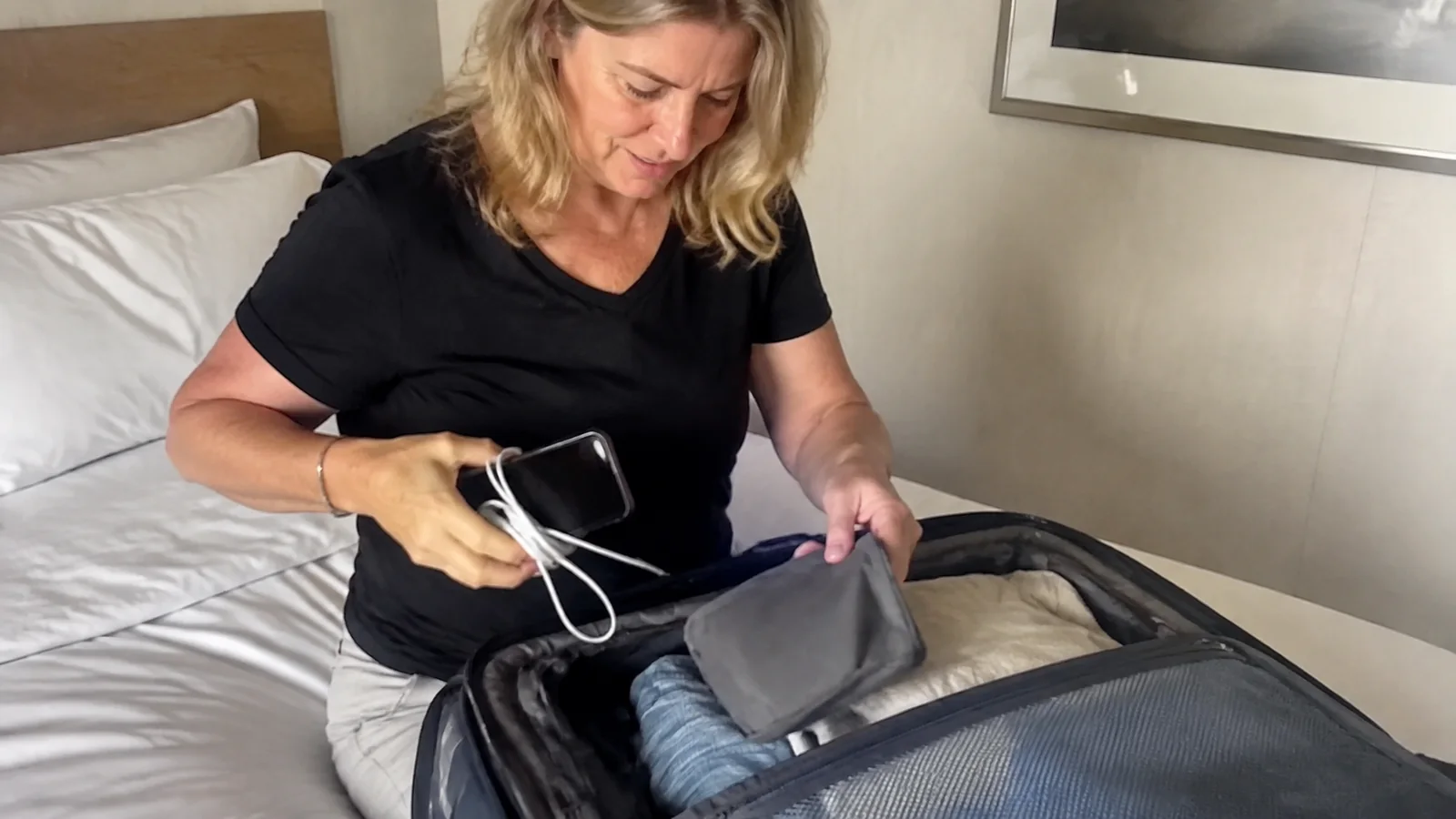 Woman packs a phone and cable into an open suitcase.