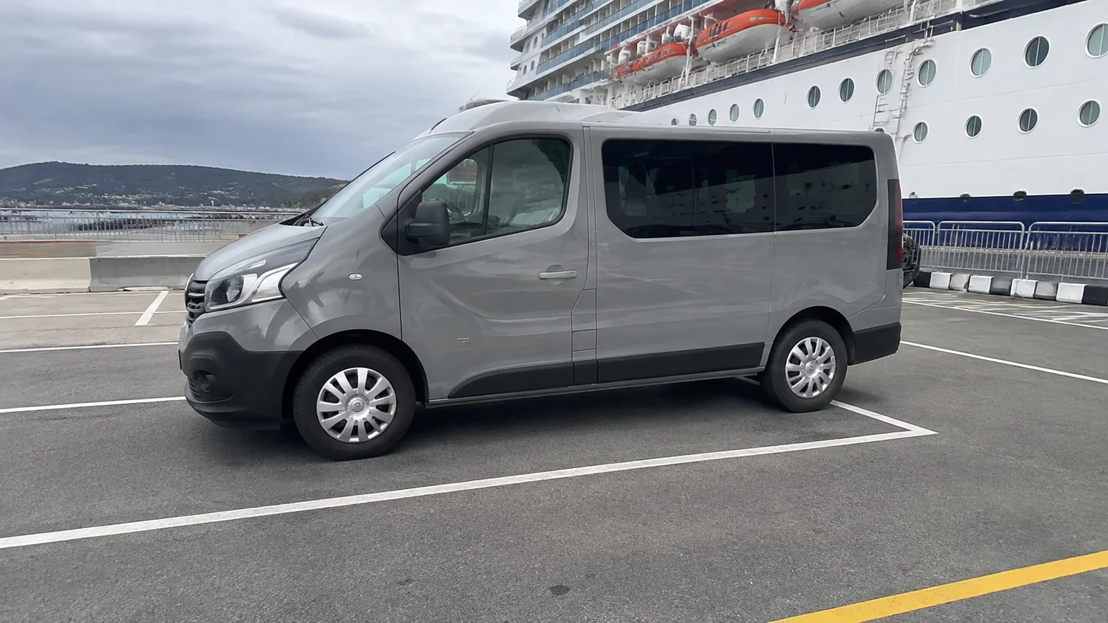 Gray passenger van is parked beside a large cruise ship.