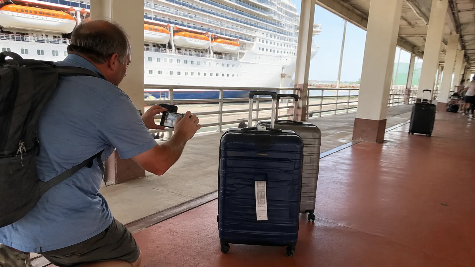 Man photographs a tagged suitcase beside a docked cruise ship.