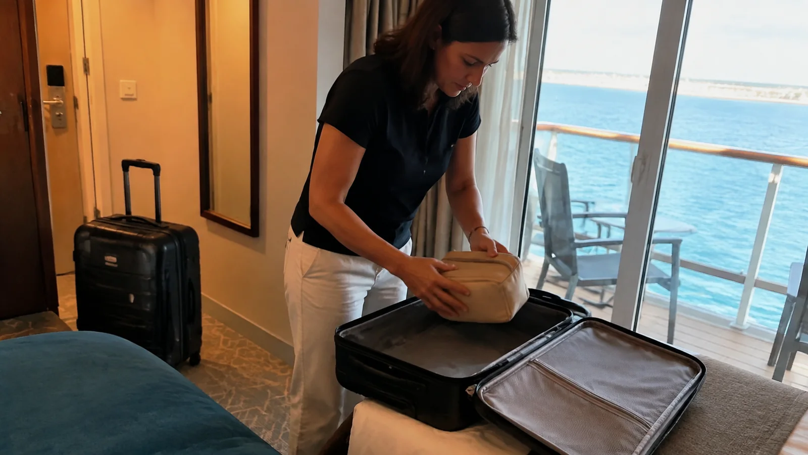 Woman packs a small pouch into a suitcase beside balcony doors.