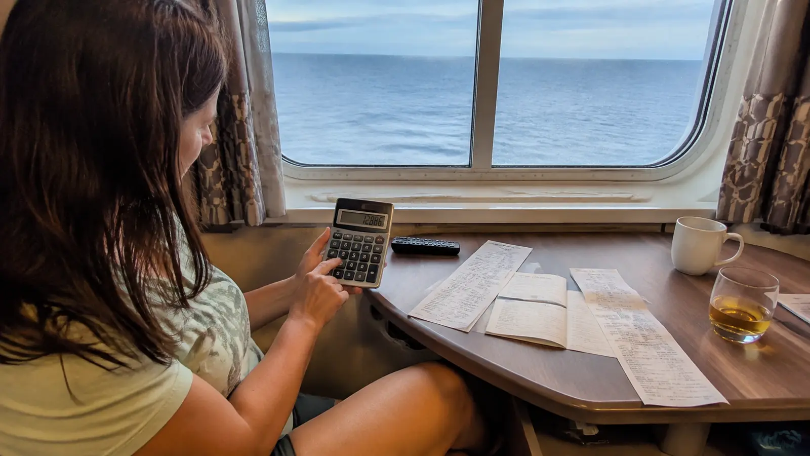 Woman using a calculator with receipts and notes at a cabin table by a large window