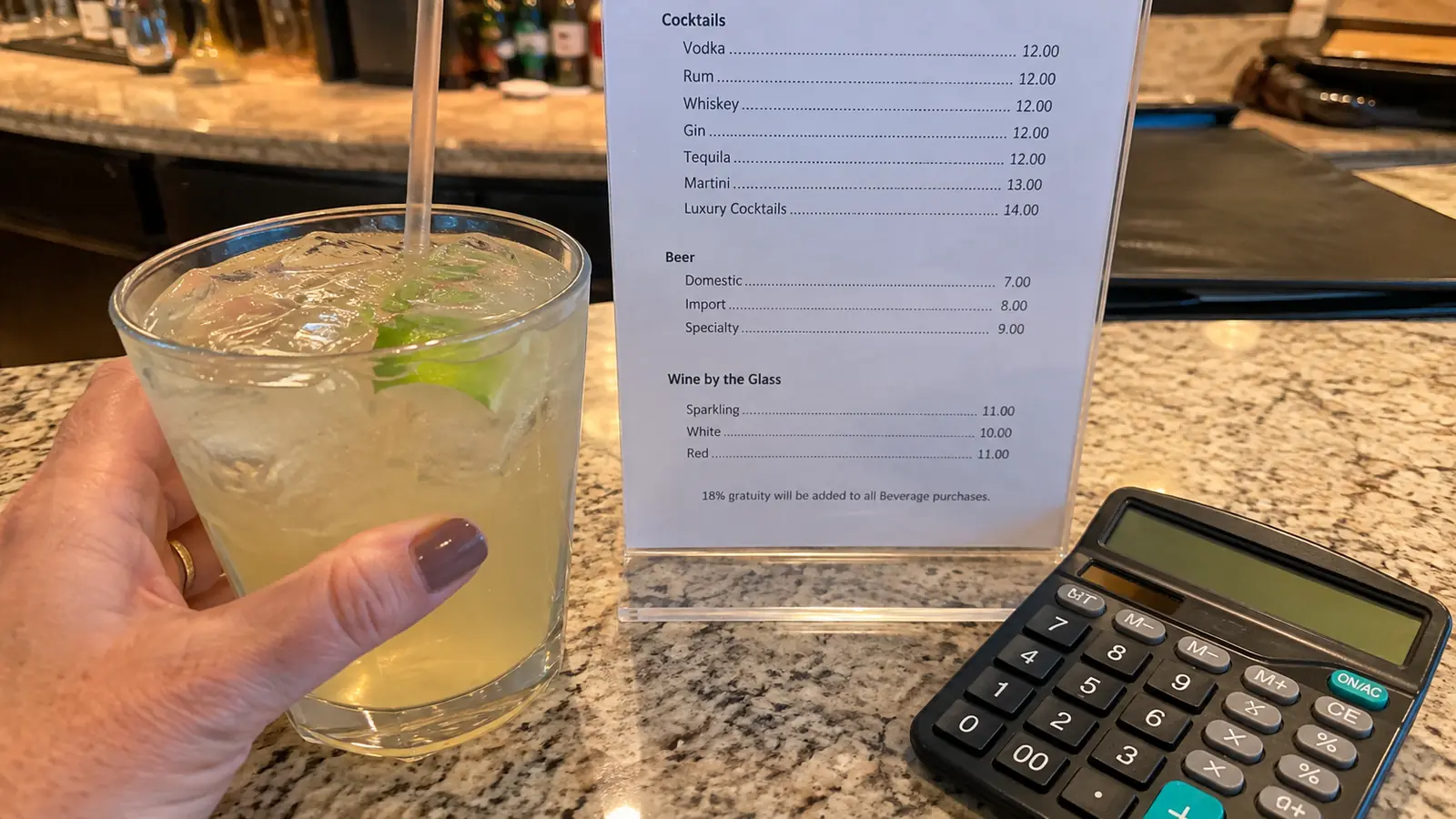 Hand holding a cocktail beside a printed drink menu and calculator at a bar