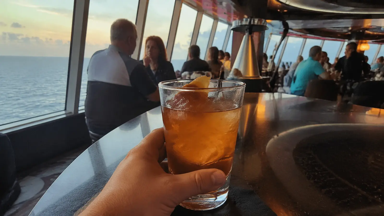 Hand holding an iced drink at a cruise ship bar with passengers and ocean views