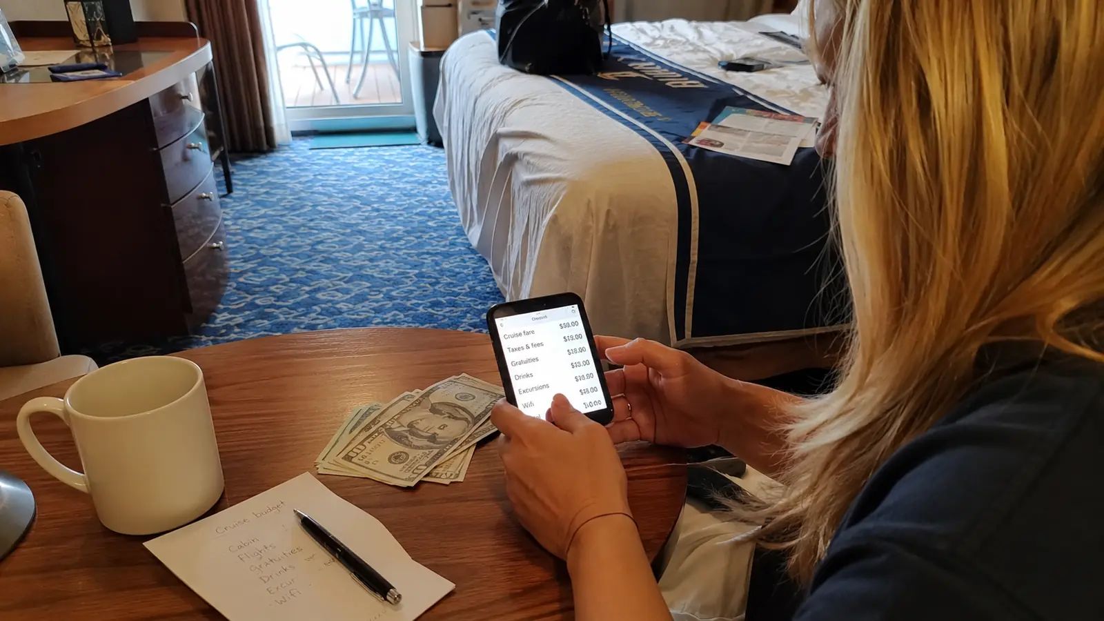 Woman checking a smartphone beside cash, a notebook, and coffee on a cabin table