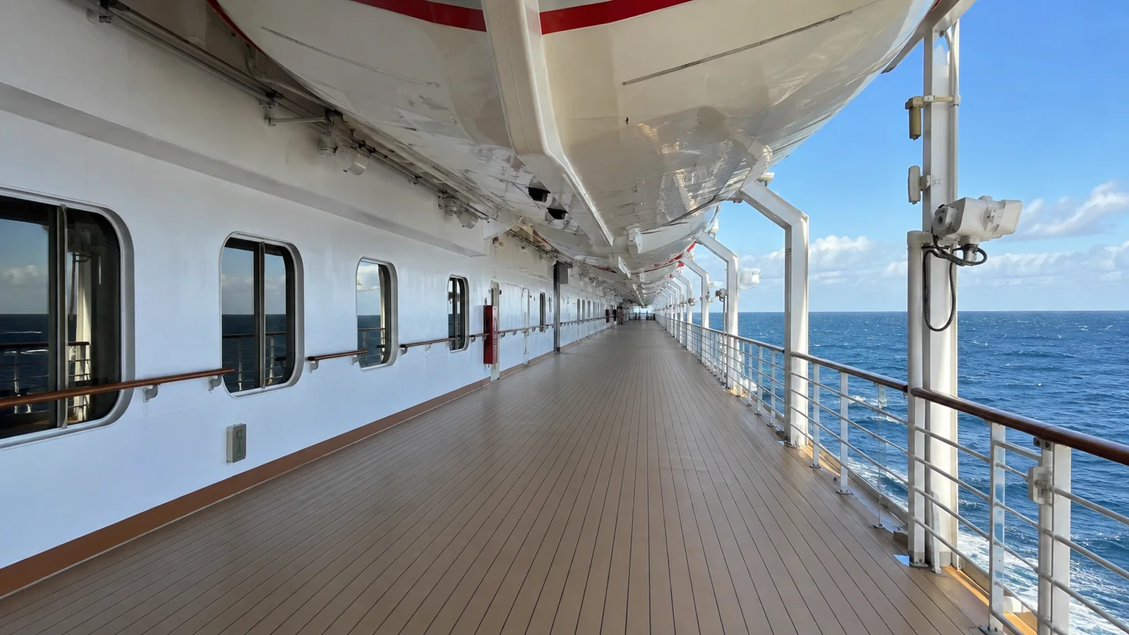 Empty exterior promenade runs beneath lifeboats beside the open sea.
