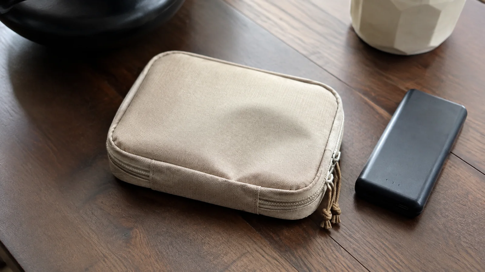 Beige zippered pouch and black rectangular device on a wooden table.