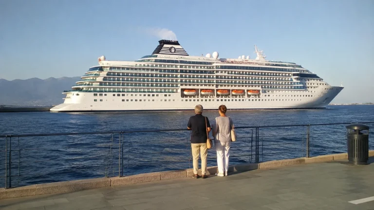 Two people standing at a waterfront railing facing a large cruise ship.