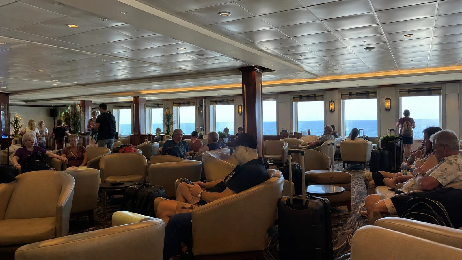 Passengers gather with their luggage in a large ship lounge.