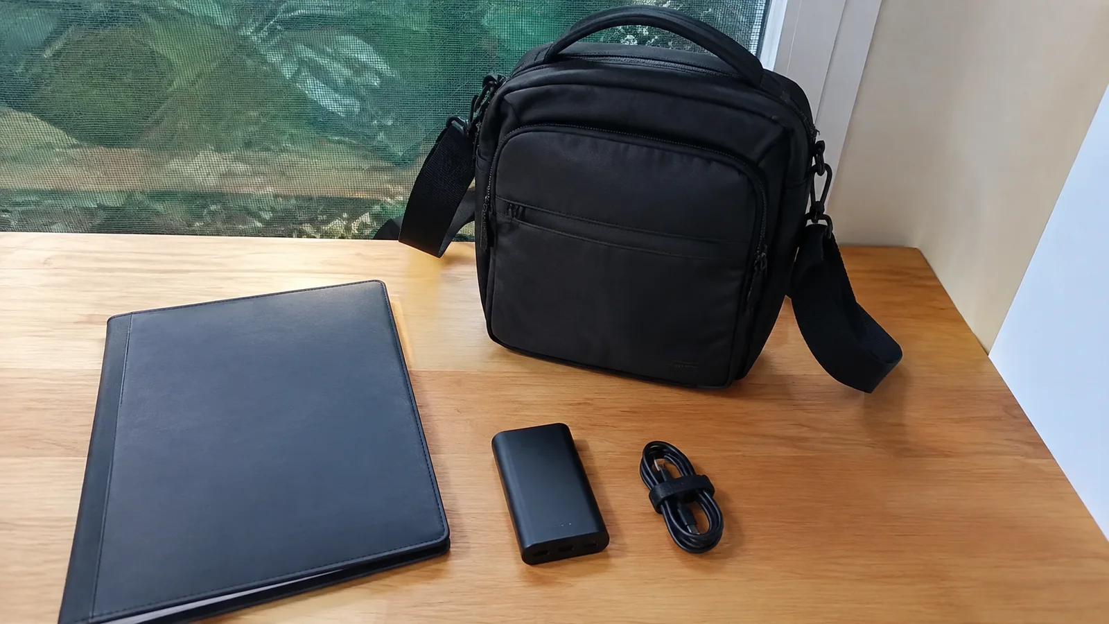 Small shoulder bag, dark folder, phone and cable lie on a table.