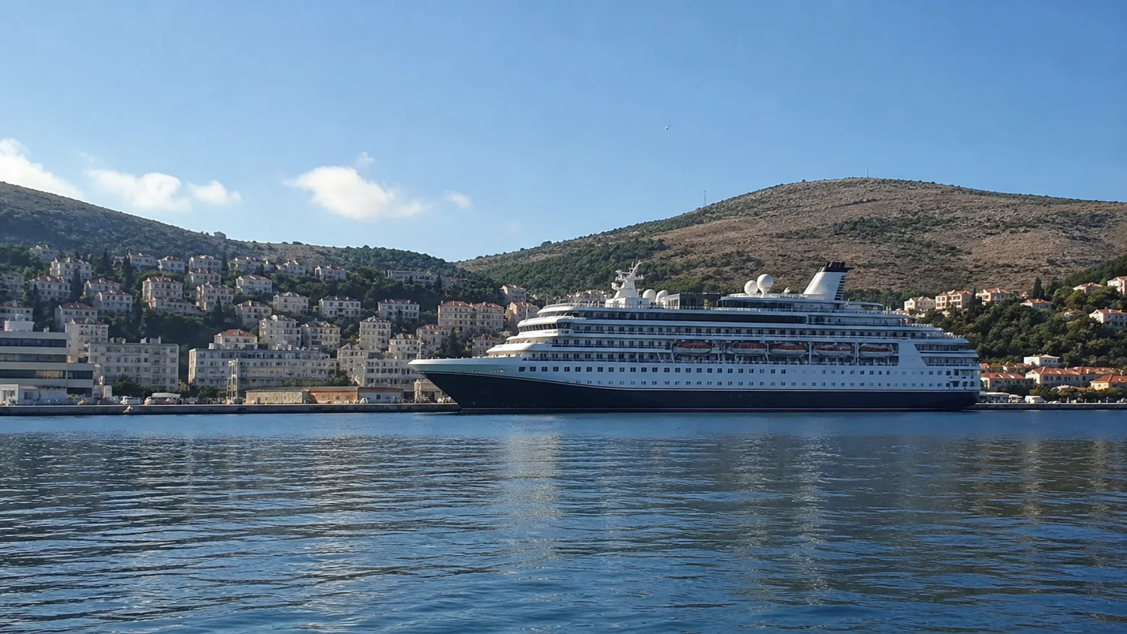 Cruise ship docked beside a hillside harbor under blue skies.